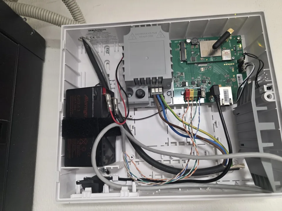 Otevřená bílá skříň ústředny s elektronikou, kabeláží a černým záložním akumulátorem.