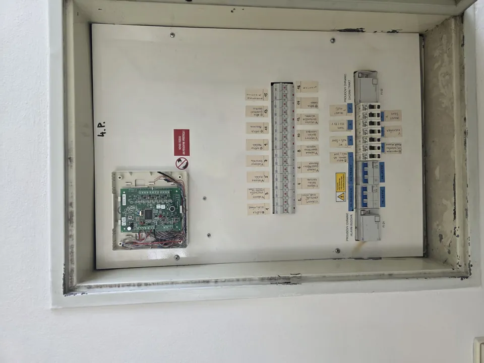 Otevřený rozvaděč s několika řadami jističů a odkrytou elektronickou deskou nahoře.