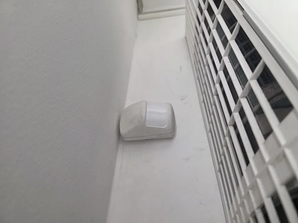 Bílý pohybový detektor v rohu místnosti nad oknem s kovovou mříží.